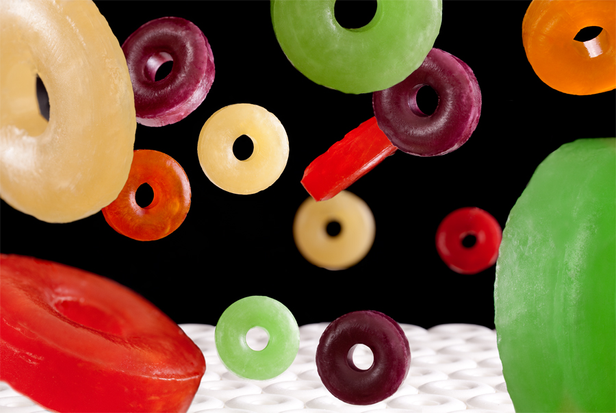 This Brian Charles Steel photograph is composed of white wheel-shaped mints. The background is black. In the bottom left corner is a red wheel-shaped candy. Above it is a yellow one. In the bottom right corner is a green wheel shaped candy. Above it is a much smaller orange candy of the same shape. In the middle are purple, yellow, red and green candies of the same shape.  